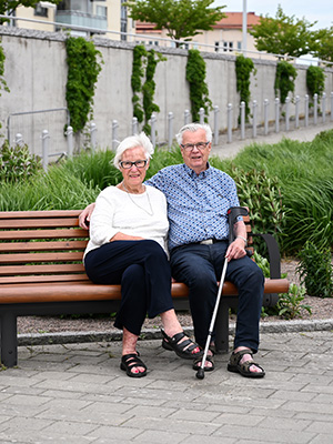 Elderly couple are sat on a lovely wooden bench with backrest, they are close together looking towards the camera.